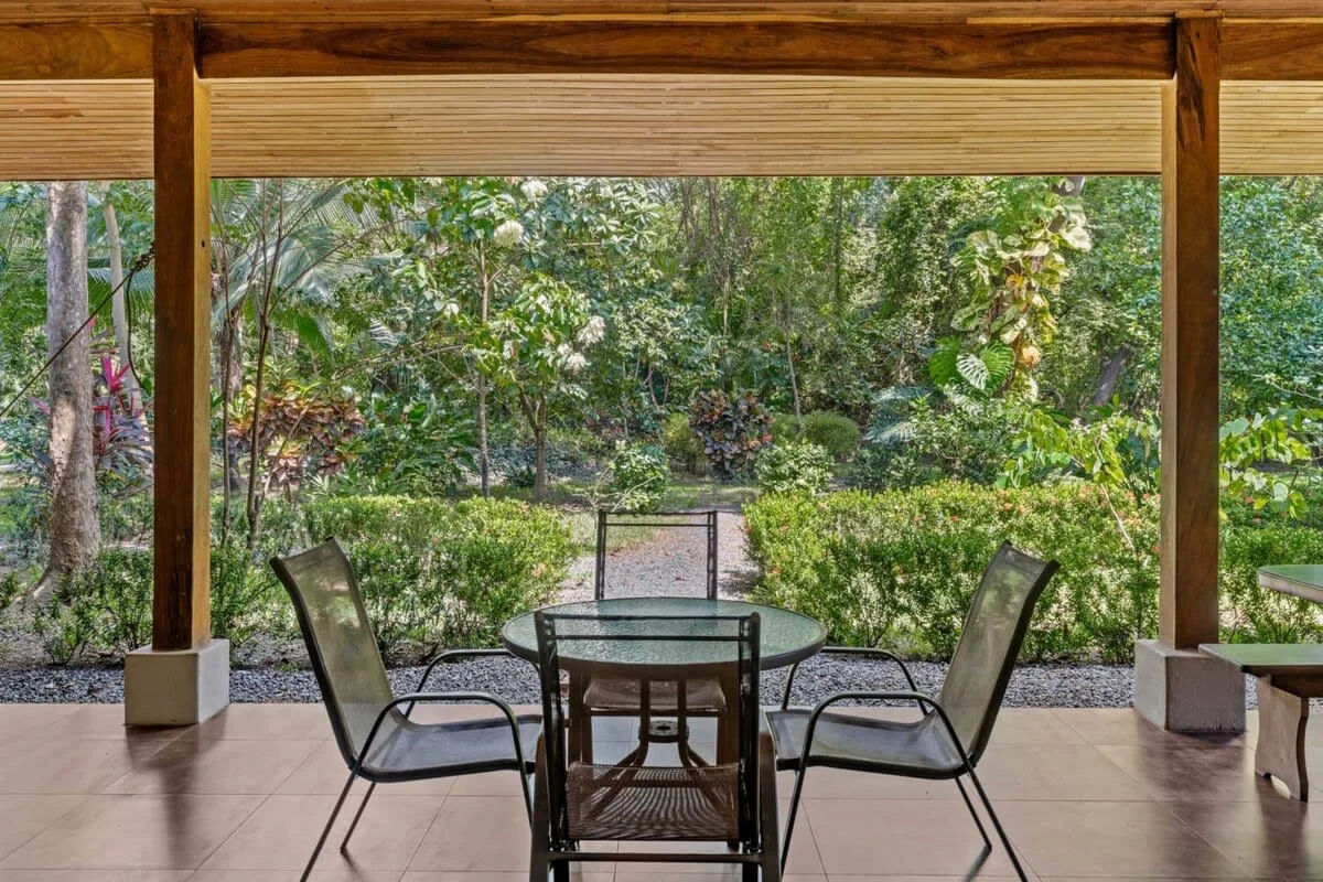 Un patio cubierto con una mesa de vidrio y asientos da a un jardín frondoso.
