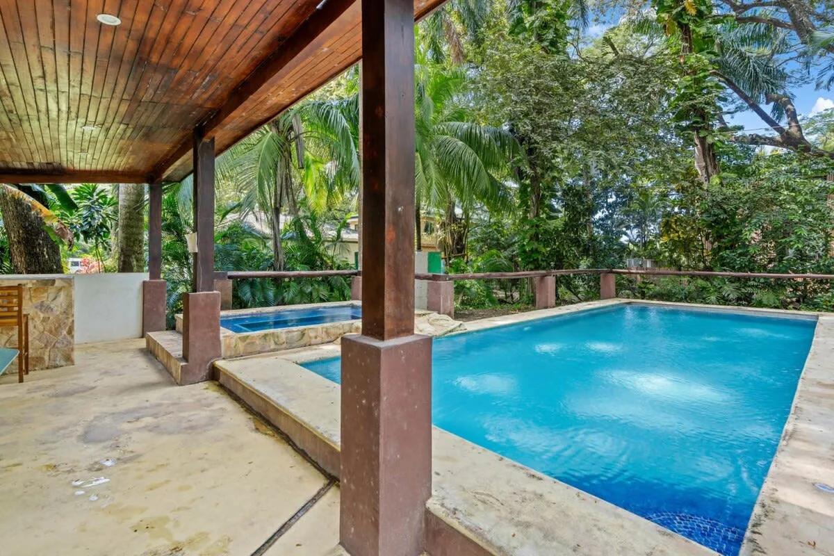 Una piscina al aire libre junto a un patio cubierto, rodeada de árboles tropicales.