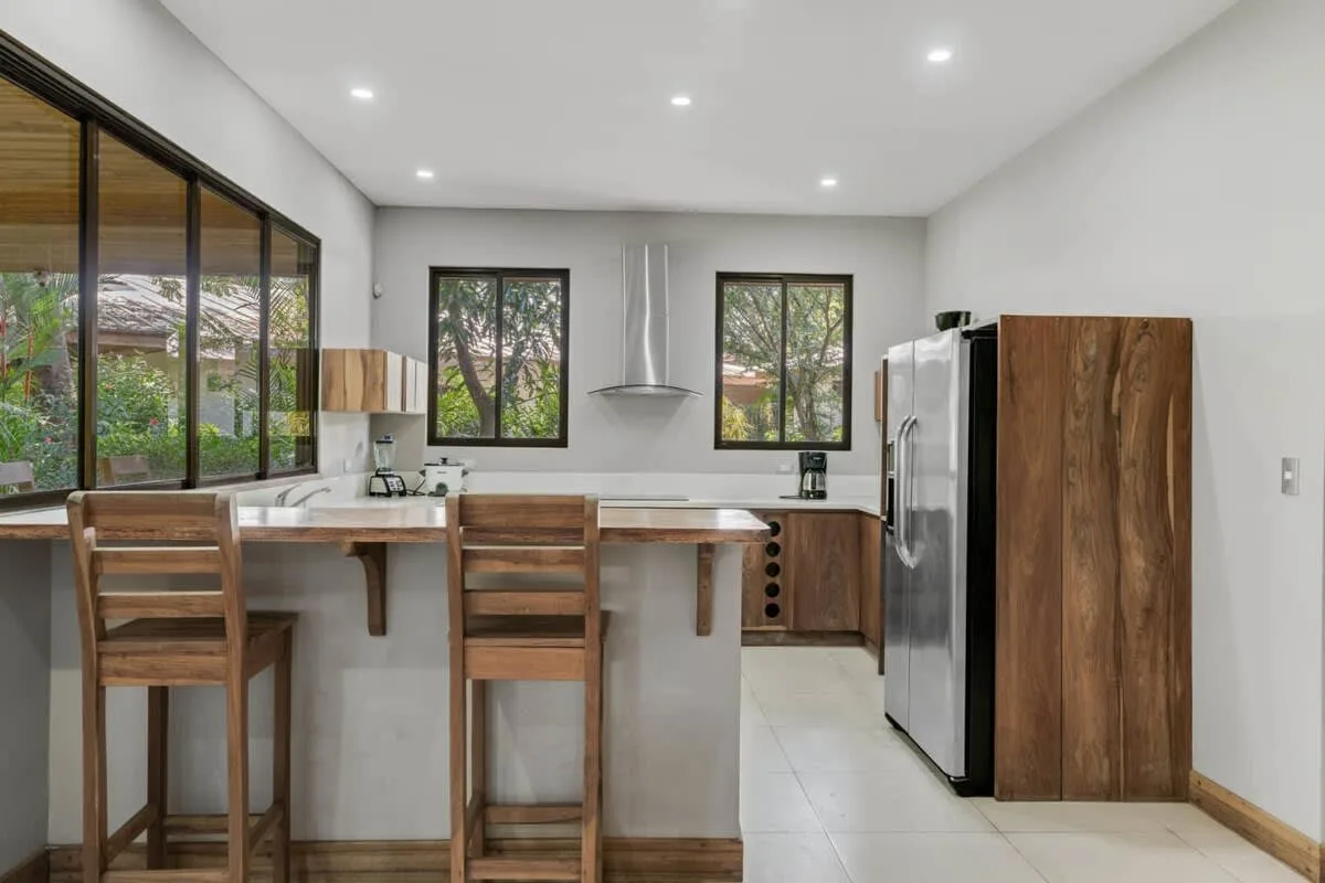 A bright kitchen with a counter, wooden stools, and large windows.
