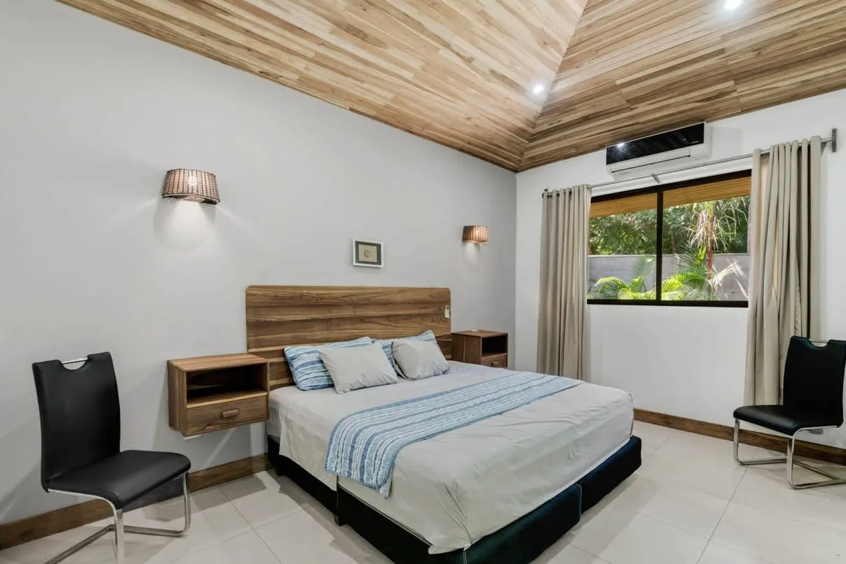 Un dormitorio con cama, mesitas de noche y una ventana con vegetación al otro lado.