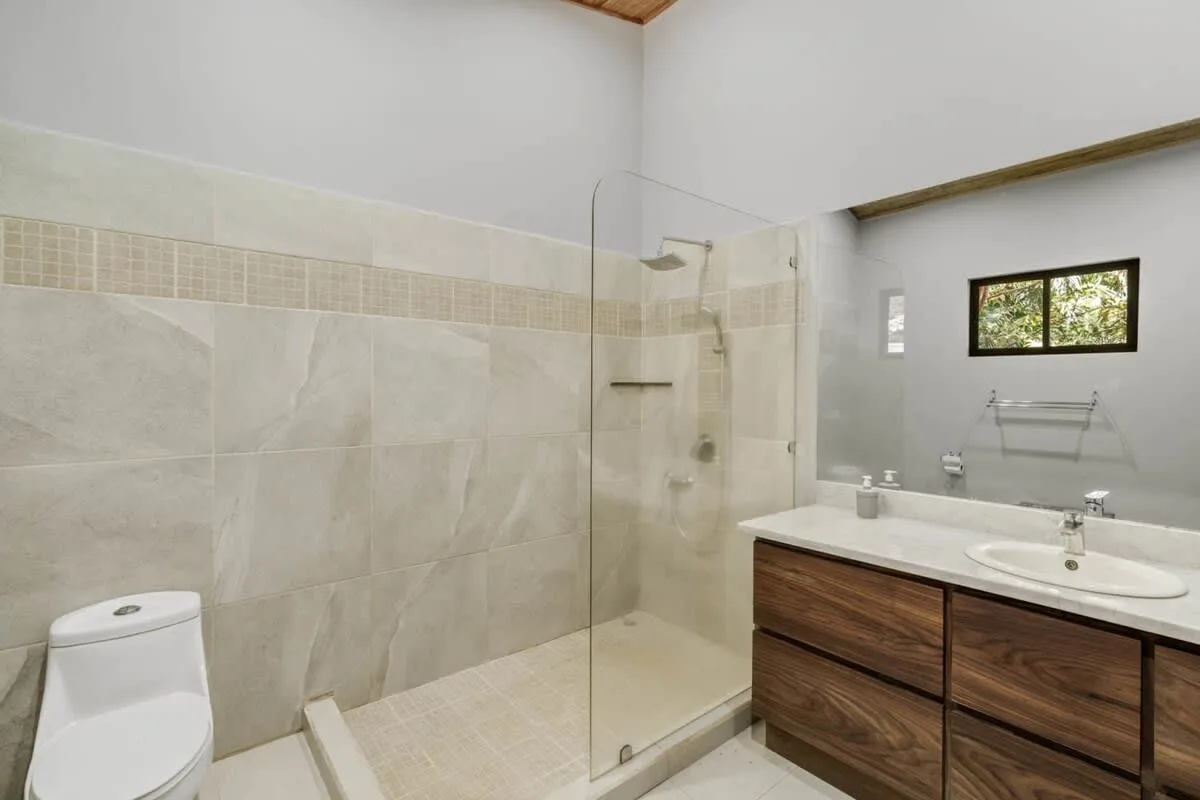 Un baño con ducha de vidrio, inodoro y tocador en tono madera.