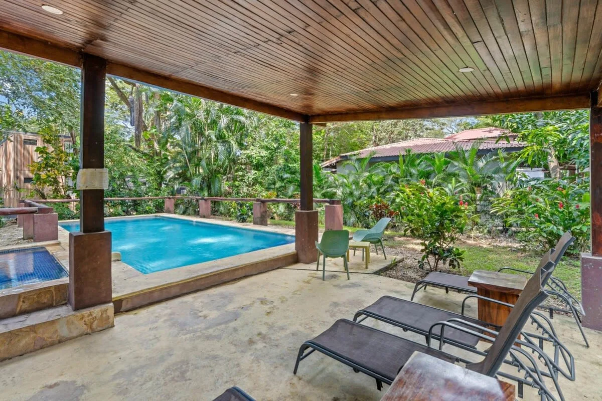 Un patio cubierto junto a la piscina tiene asientos y vistas a un jardín frondoso.