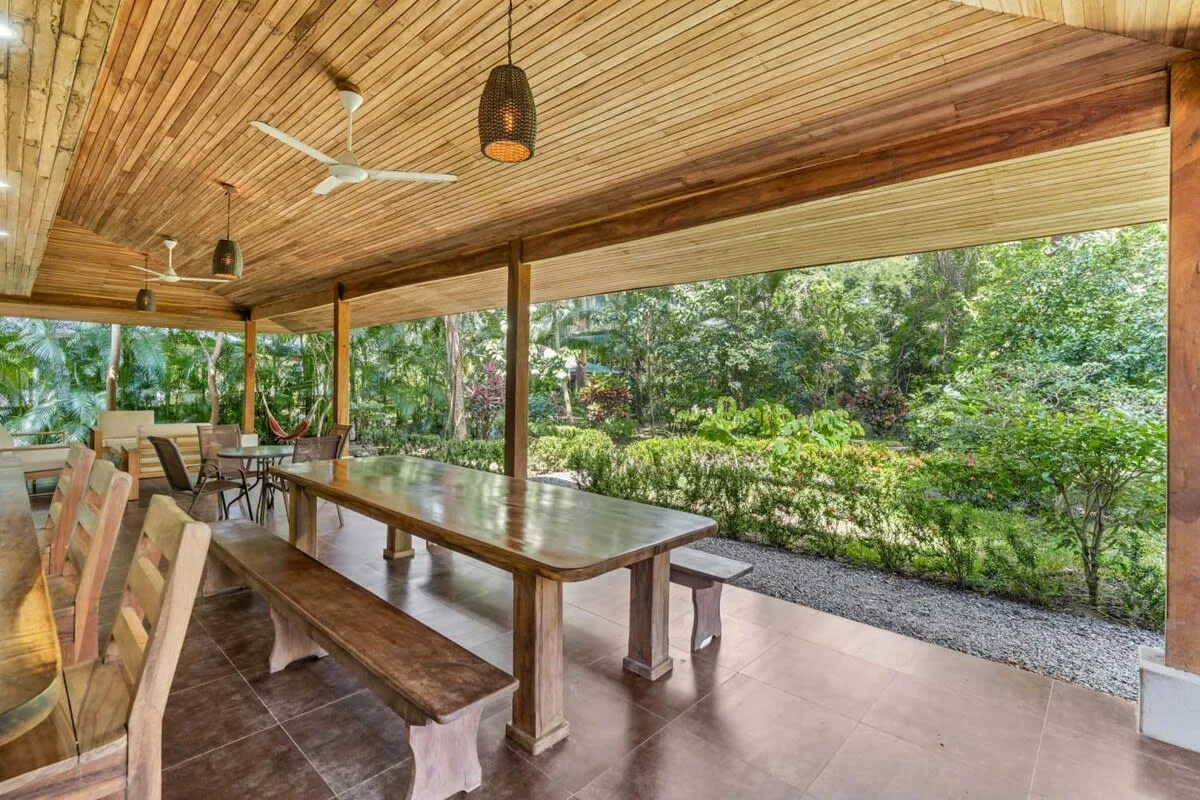Un comedor exterior cubierto con una larga mesa de madera y vistas al jardín.