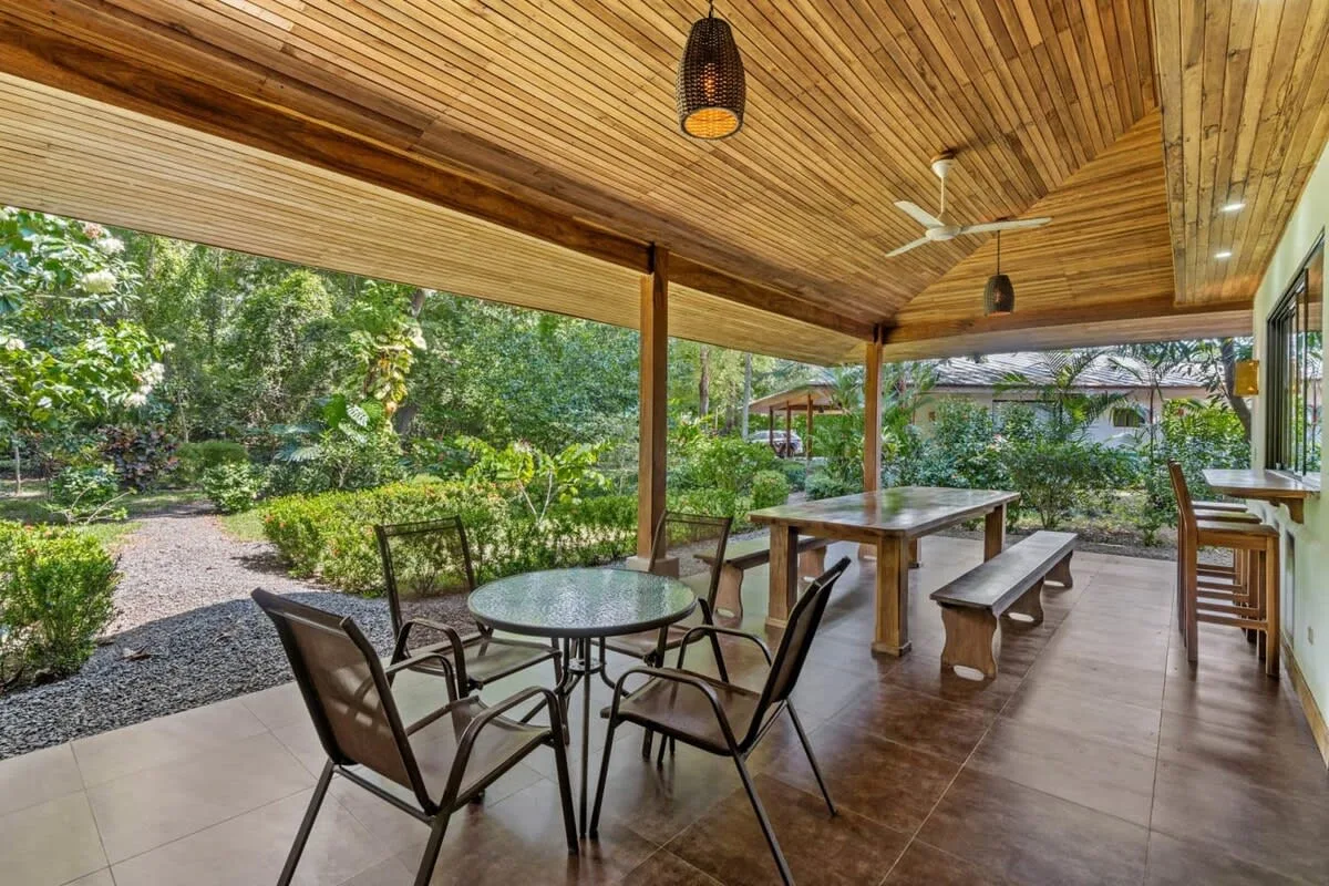 Un comedor exterior cubierto con mesas y asientos da a un jardín verde.