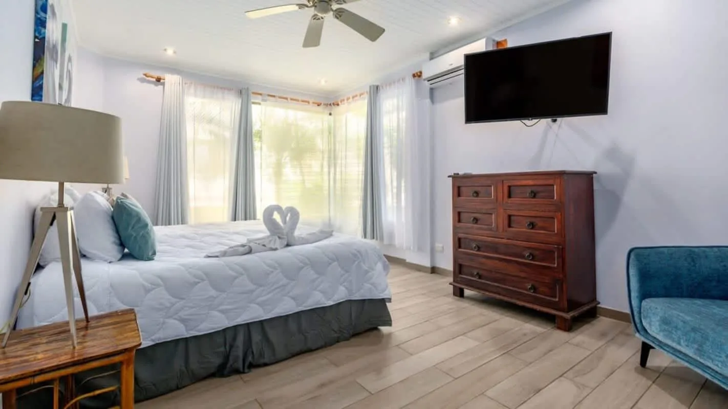 A bedroom with a bed, dresser, and television beside bright windows.