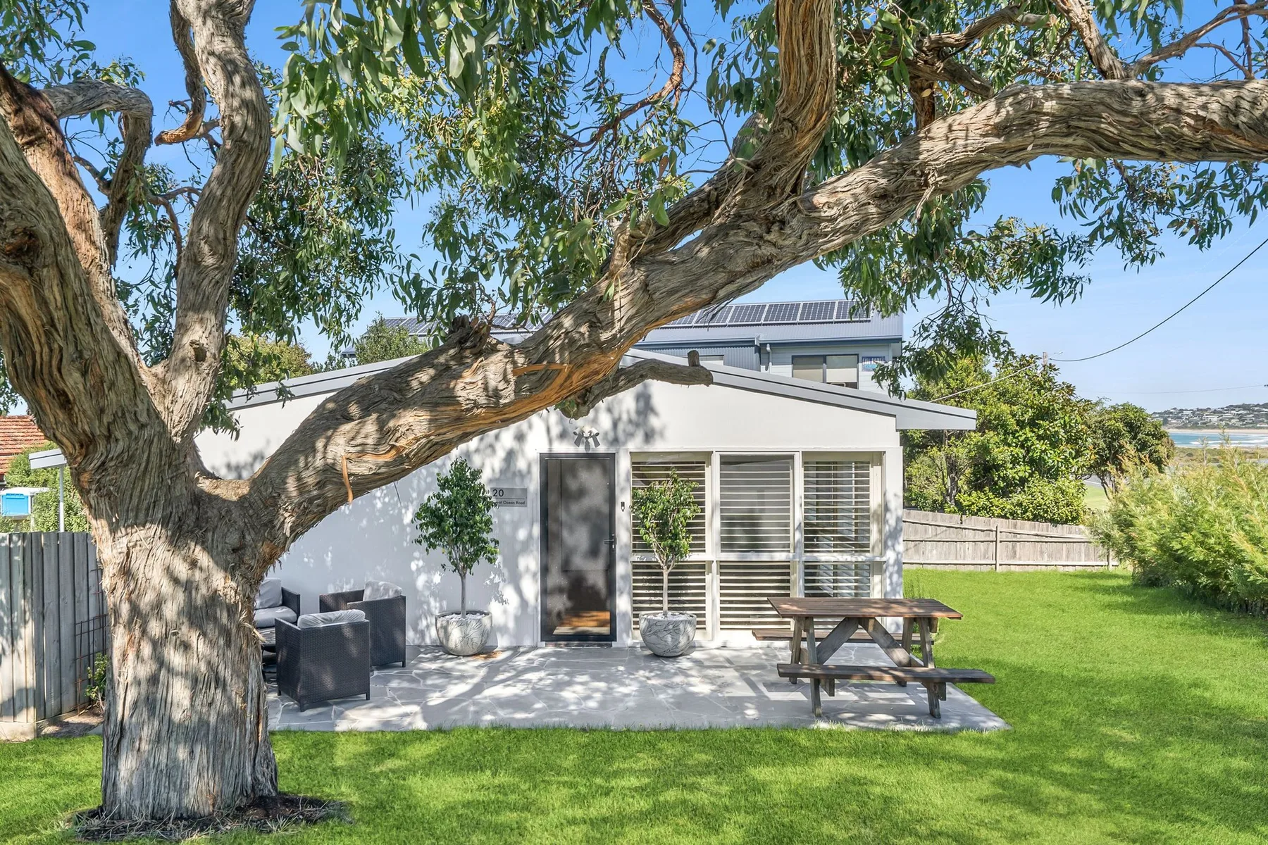 A small house opens onto a patio with outdoor seating beneath a large tree.
