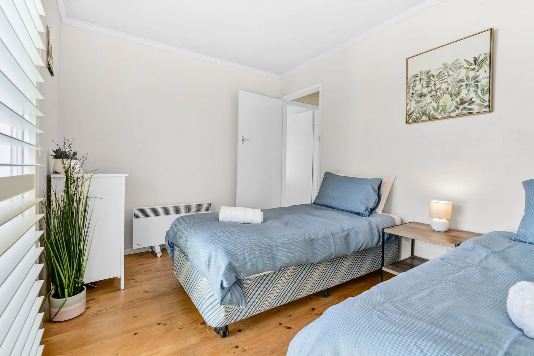 A bright bedroom with two beds, a framed botanical print, and a small bedside table.