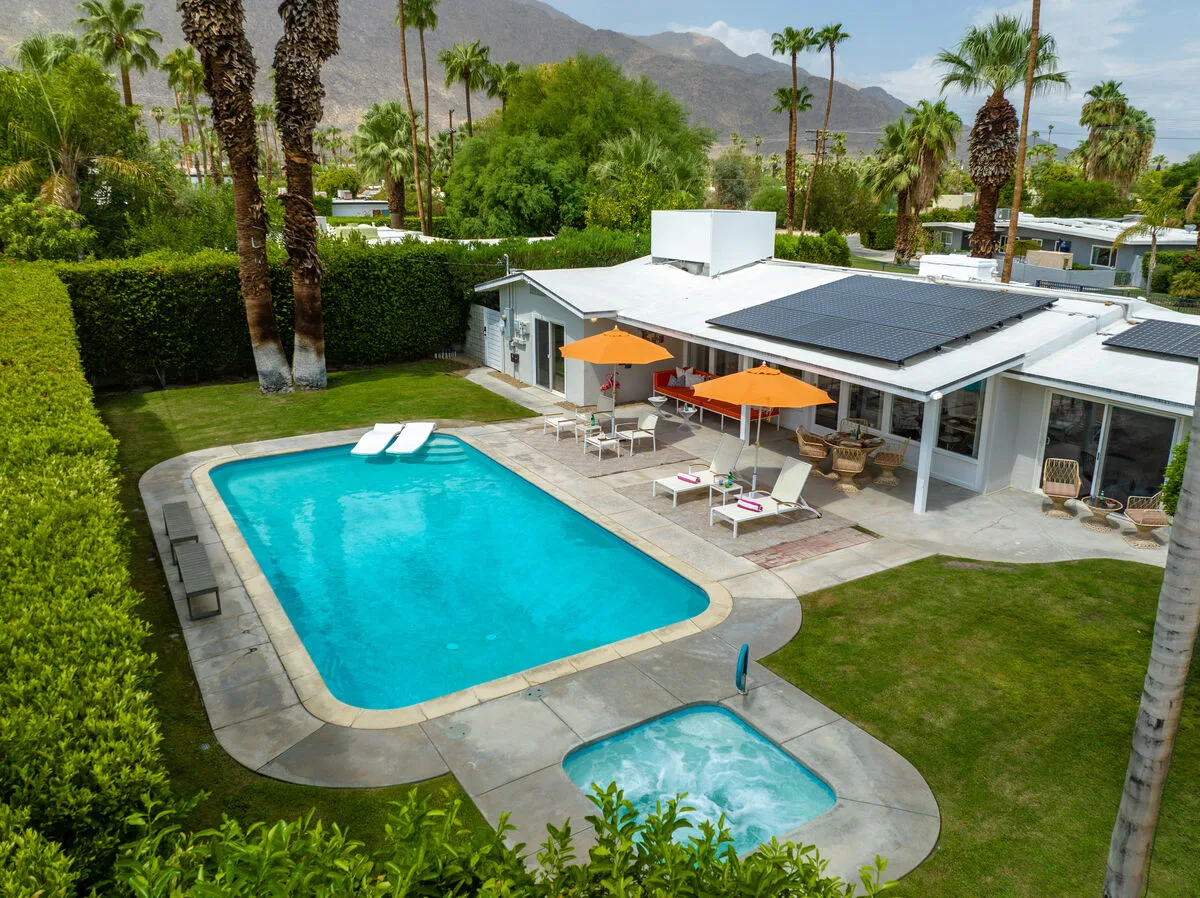 A backyard pool beside a low modern house, with patio seating and palm trees.