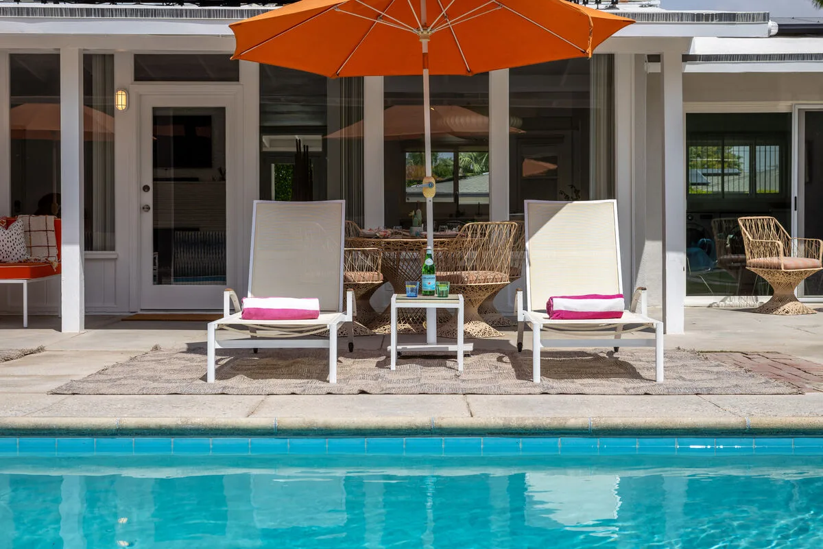 Poolside seating sits beneath an orange umbrella beside a glass-walled patio.