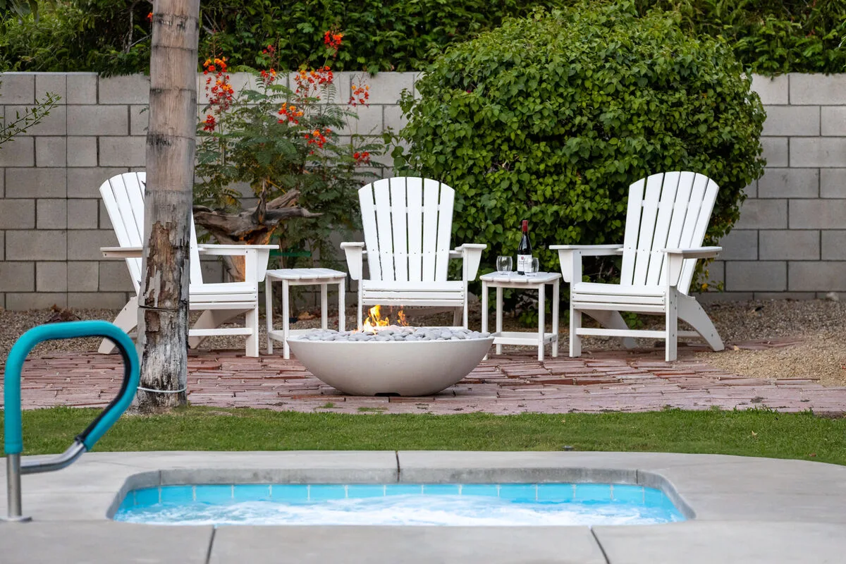 A garden patio with white seating surrounds a small fire pit beside a pool.
