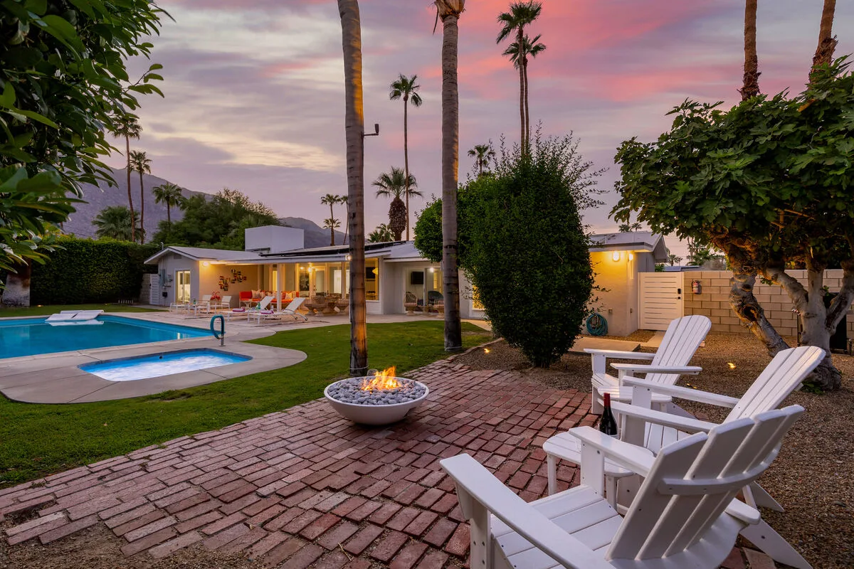 A swimming pool and patio seating sit beside a low house beneath a colorful evening sky.