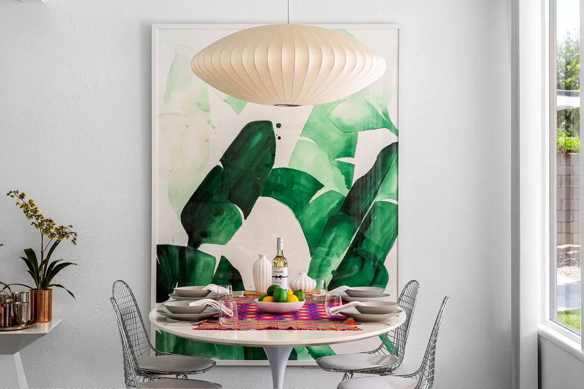 A dining area with a round table, wire seating, and large botanical wall art.