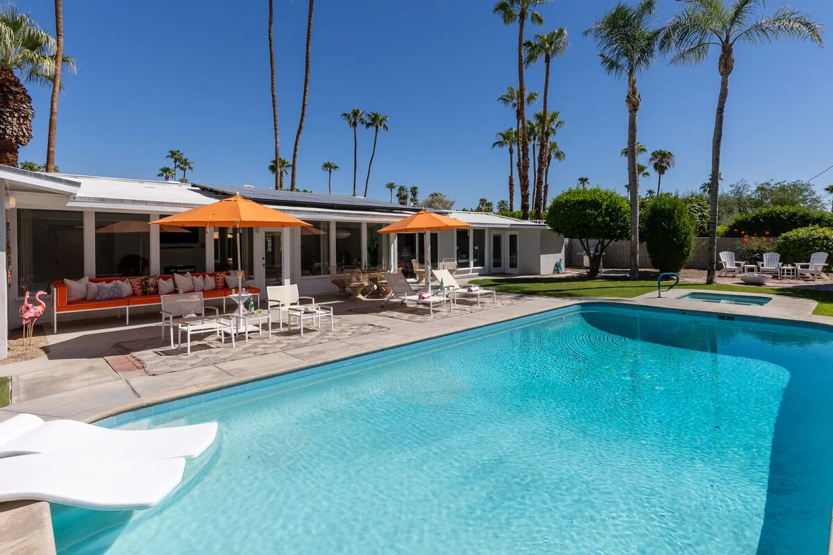 An outdoor swimming pool beside a low house, with patio seating and orange umbrellas.