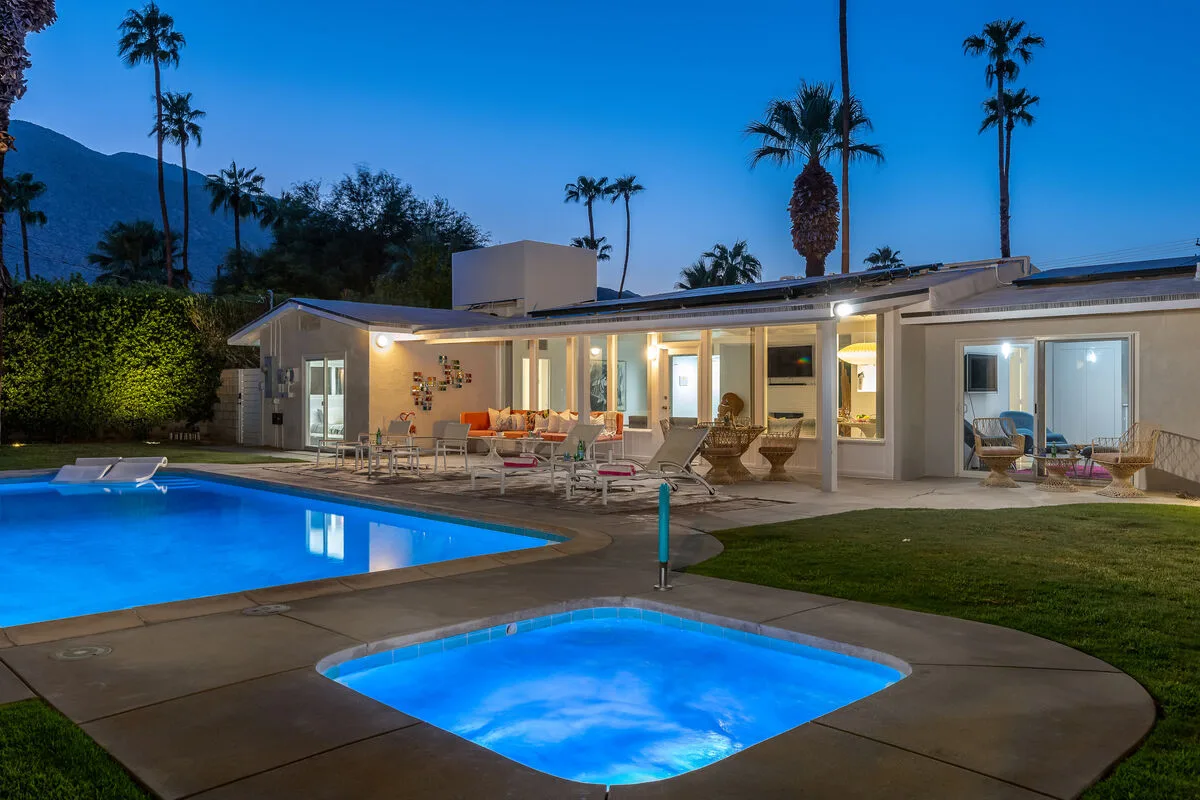 A modern house opens onto a pool and smaller illuminated spa at dusk.