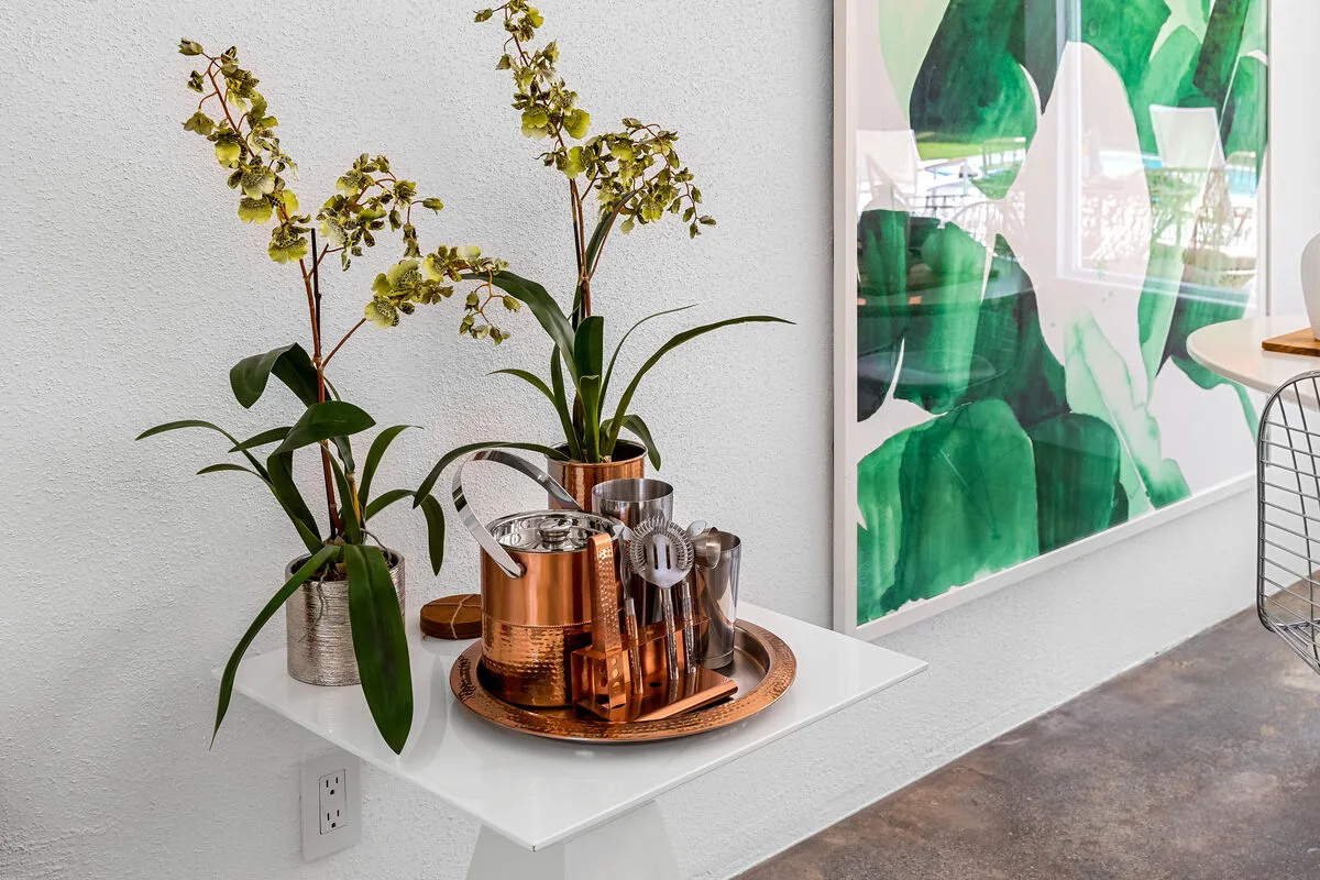 A white side table displays flowering plants and a copper drink tray beside a large green artwork.