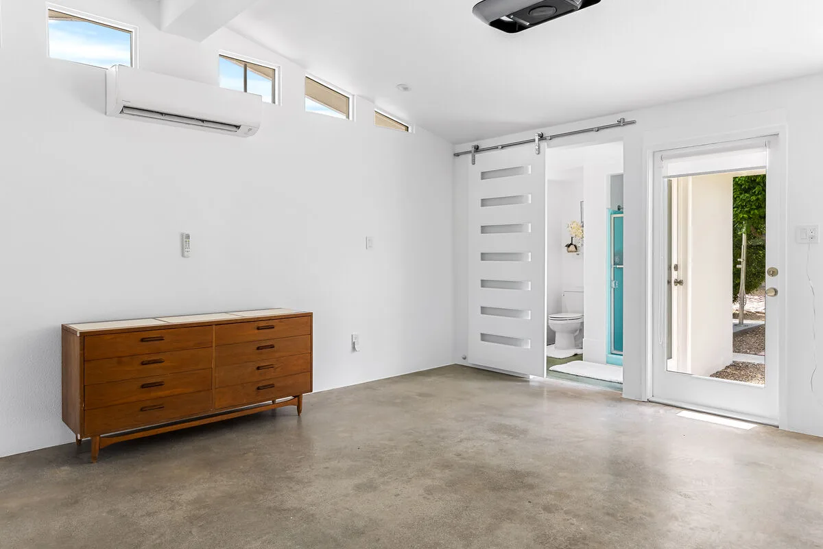 A bright, white room has a wooden dresser and an open bathroom doorway.