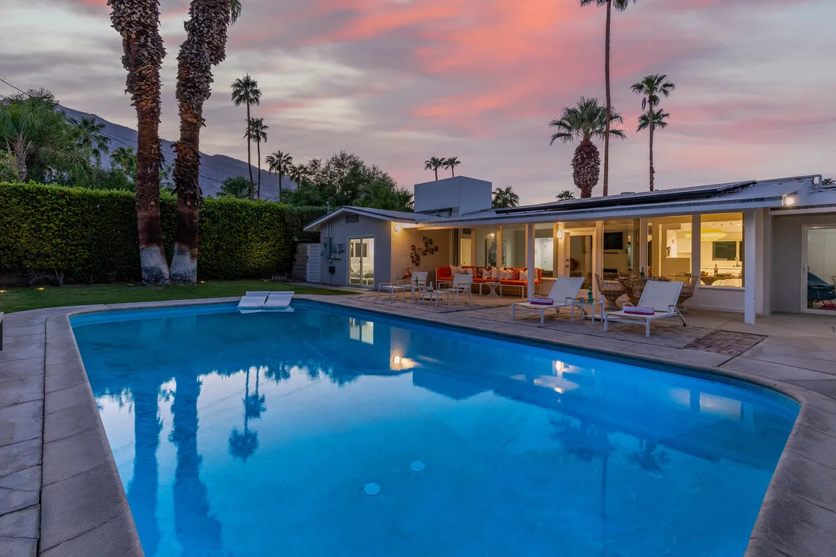A swimming pool beside a modern house, with palm trees under a pink sunset sky.