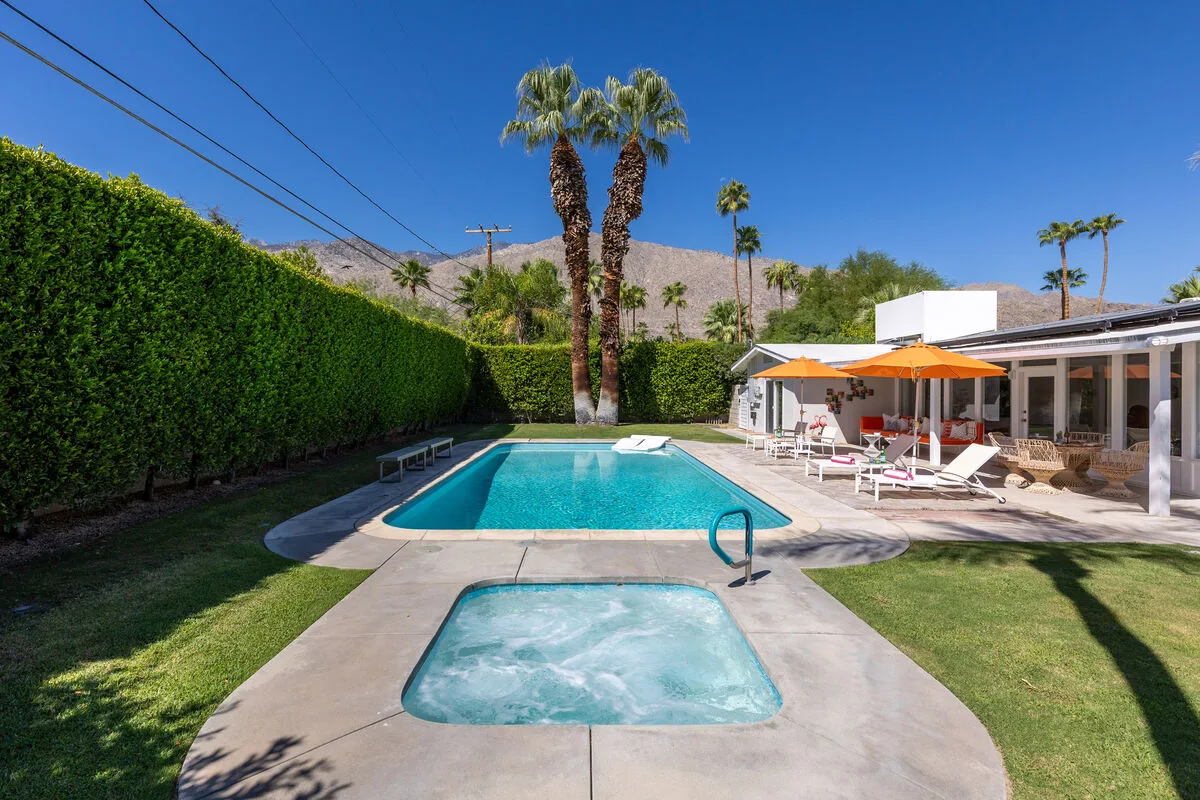 A backyard pool and spa sit beside a patio with lounge seating and an orange umbrella.