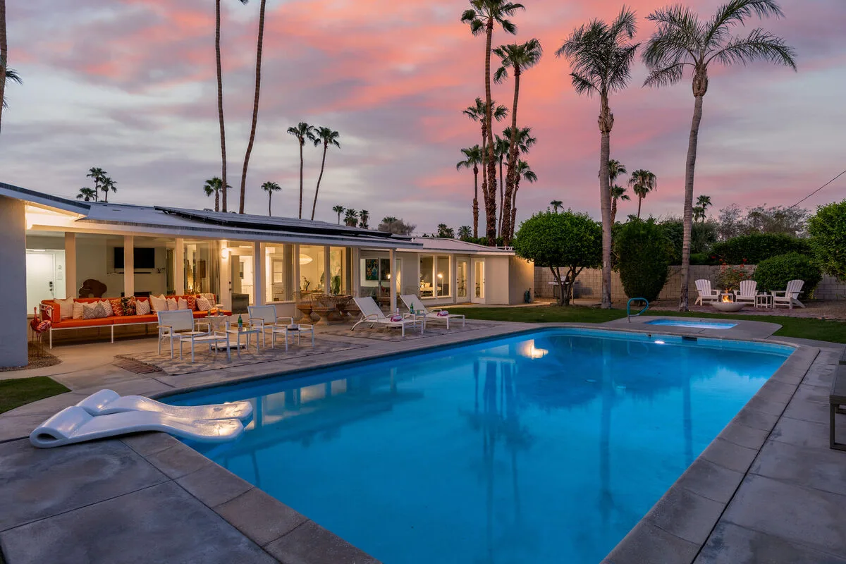 Outdoor swimming pool beside a low modern house, with palm trees under a colorful evening sky.