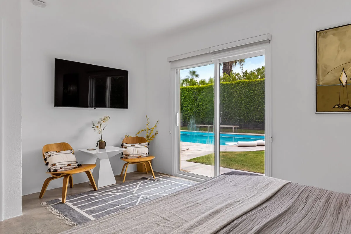 A bright bedroom with a bed, two chairs, and sliding glass doors overlooking a pool.