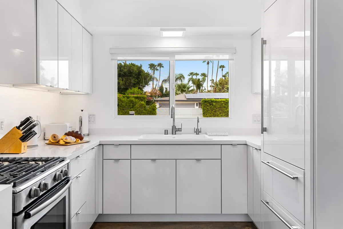 A bright white kitchen with a central window overlooking trees and palm trees.