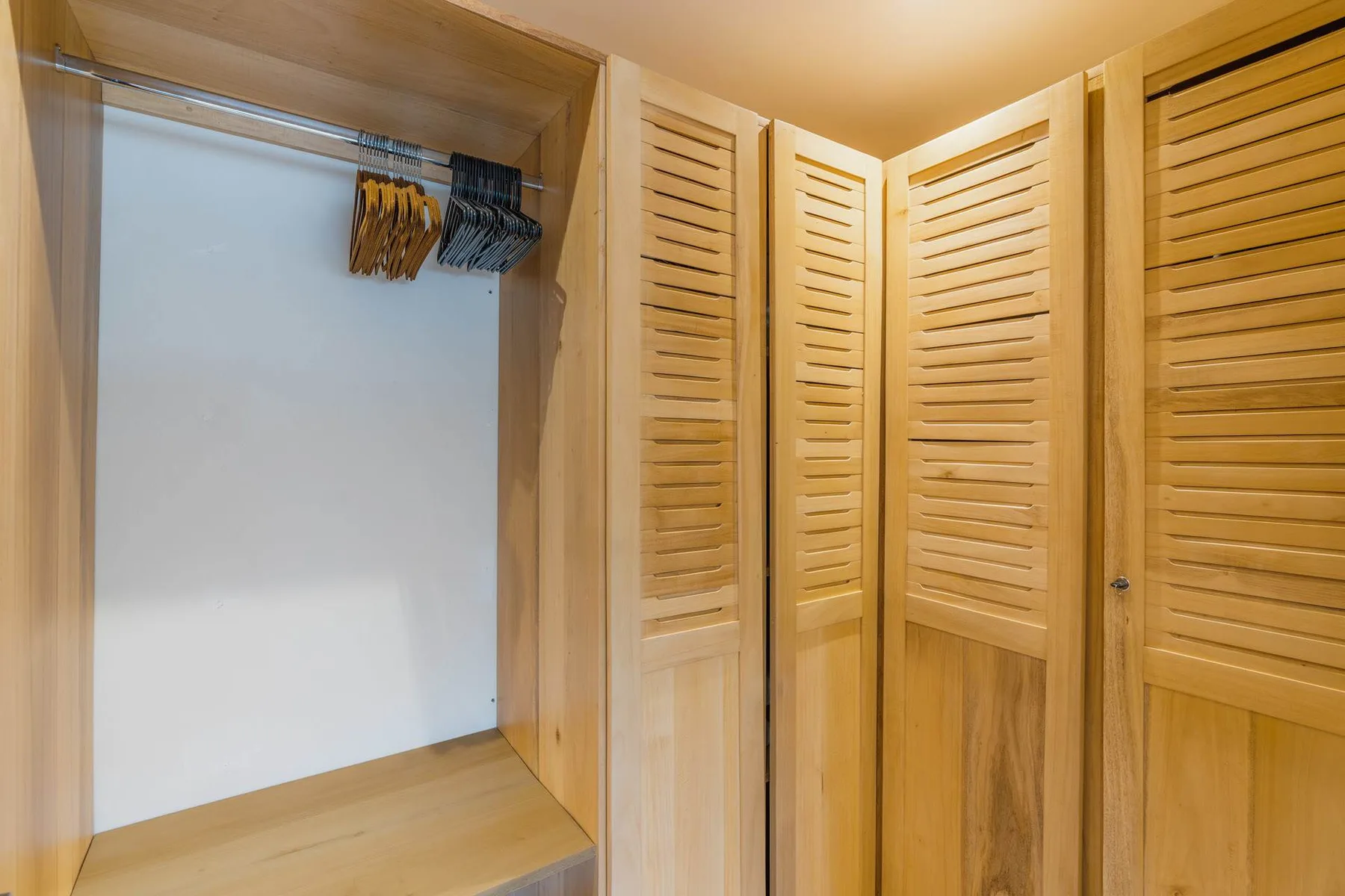 A wooden closet area with hanging clothes and louvered doors.