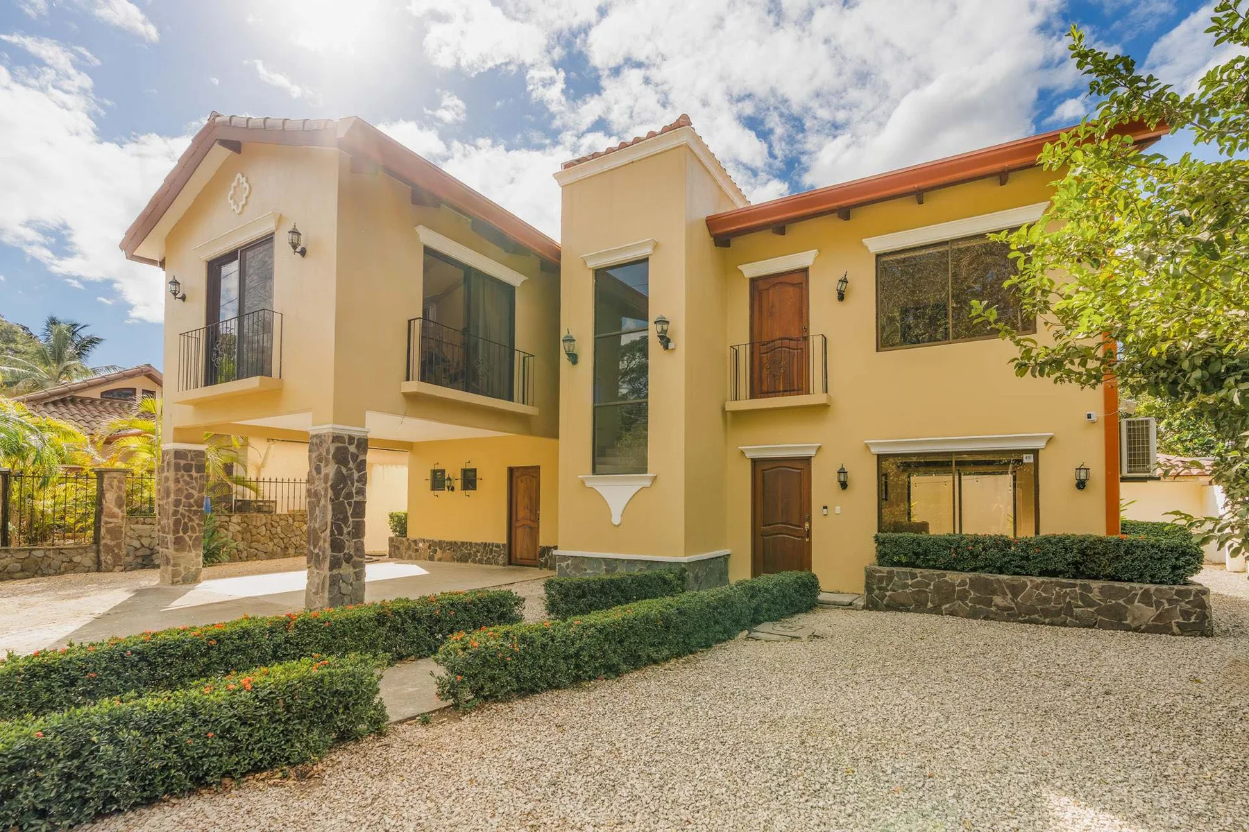 A two-story yellow house with balconies and trimmed hedges along the gravel driveway.