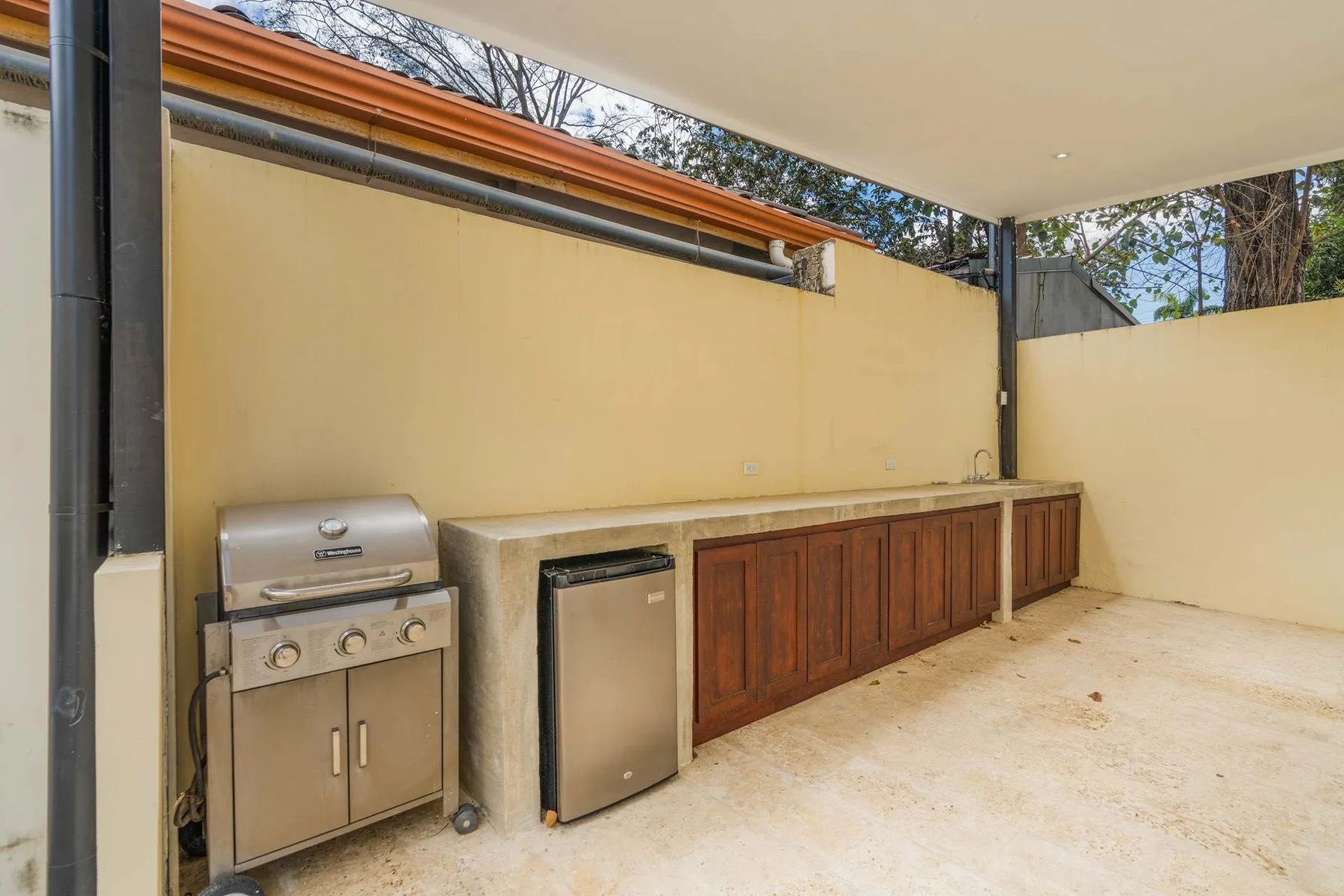 A covered outdoor kitchen with a grill, refrigerator, and wood-fronted counter.