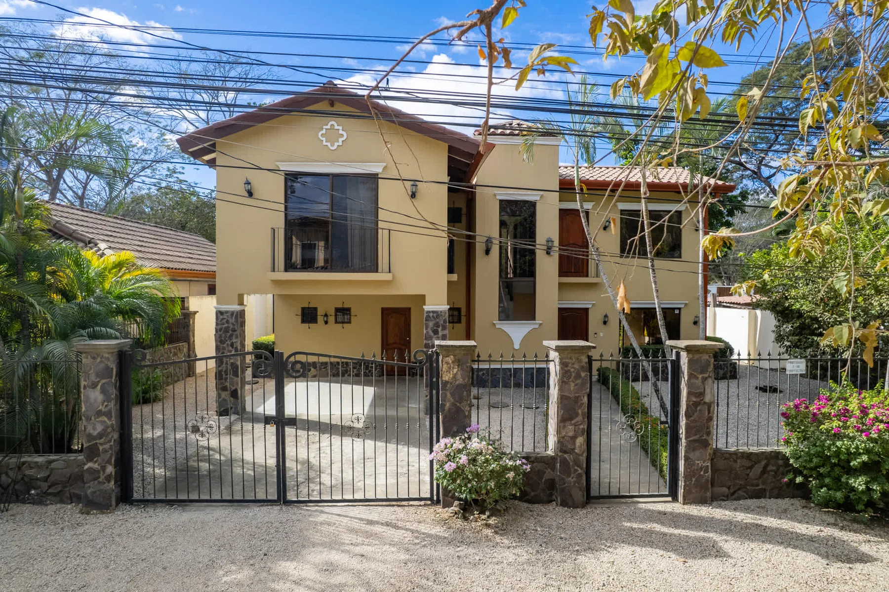 A two-story house stands behind a metal gate, with trees and greenery around it.