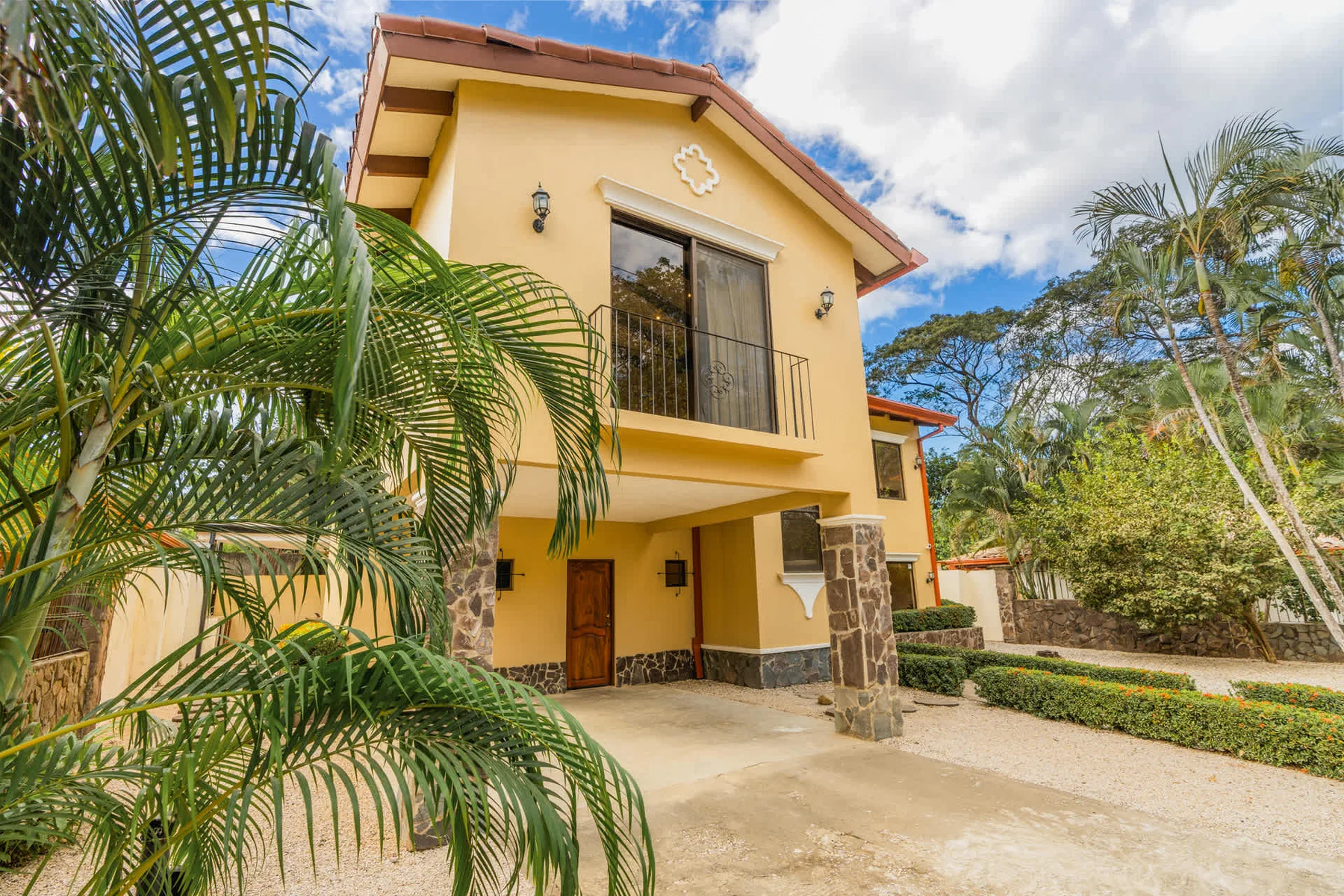A yellow two-story house stands amid palm trees beside a paved driveway.