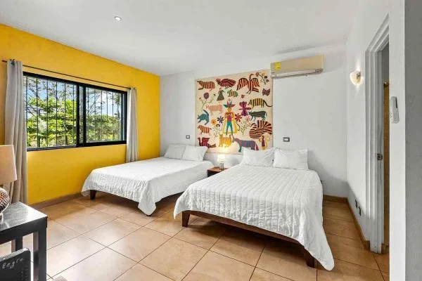 Un dormitorio luminoso tiene dos camas, una pared amarilla y una ventana grande con rejas negras.