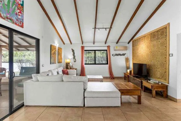 Una sala luminosa tiene un sofá seccional blanco, una mesa de centro de madera y un televisor bajo vigas expuestas.