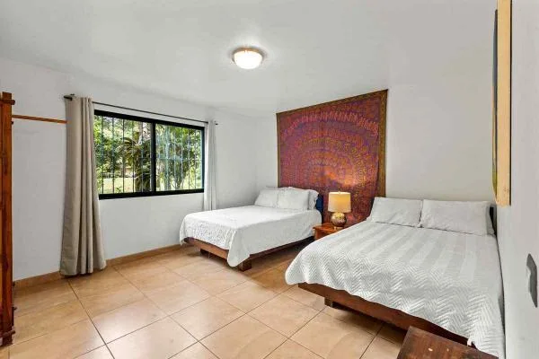 Un dormitorio luminoso tiene dos camas, una ventana con rejas y cortinas, y un tapiz colorido.