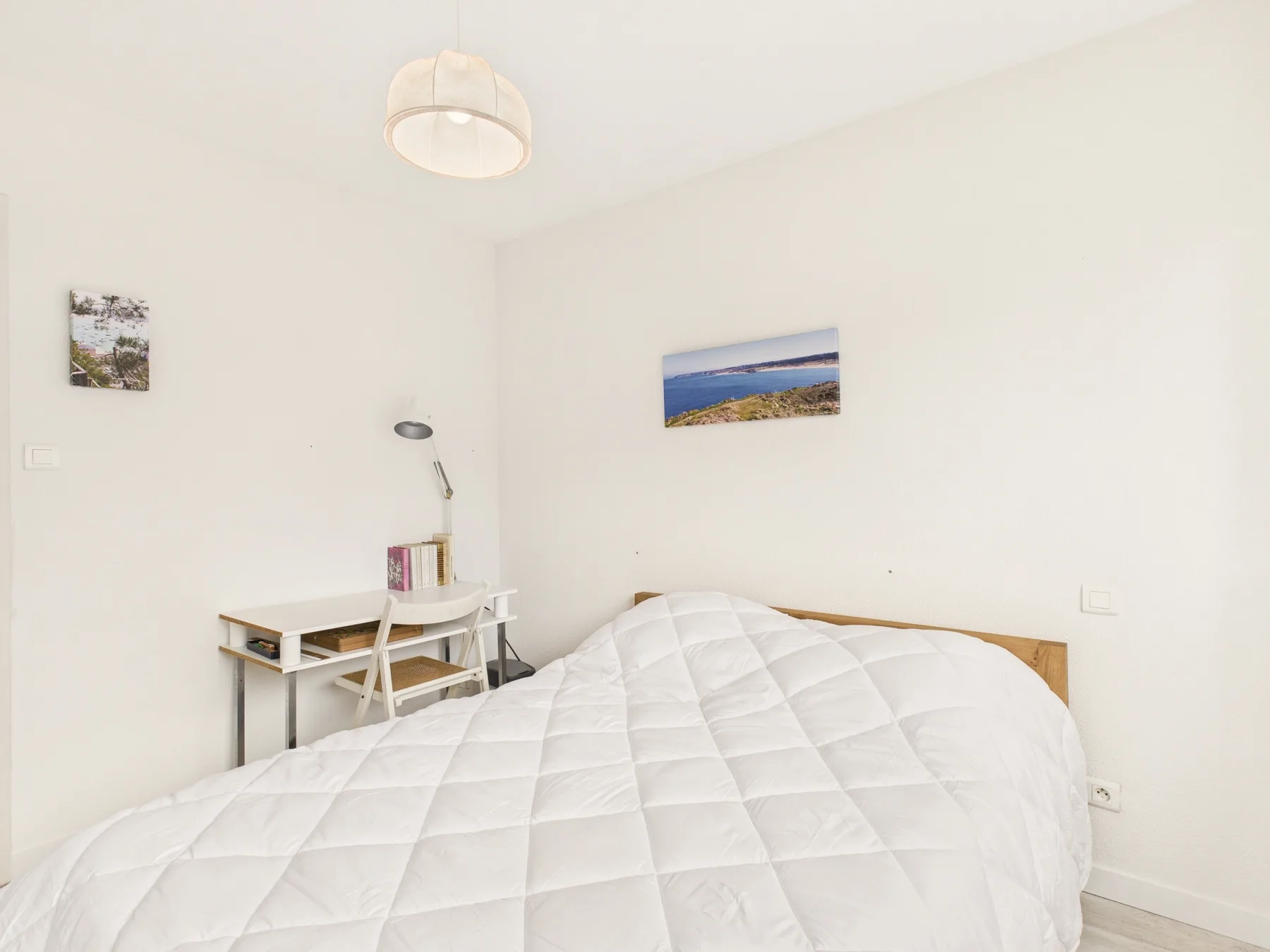 Un dormitorio luminoso con una cama blanca y un pequeño escritorio junto a la pared.