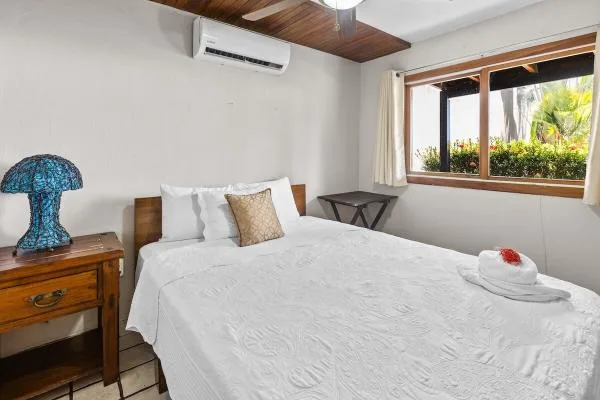 Un dormitorio con una cama blanca junto a una ventana con vegetación.