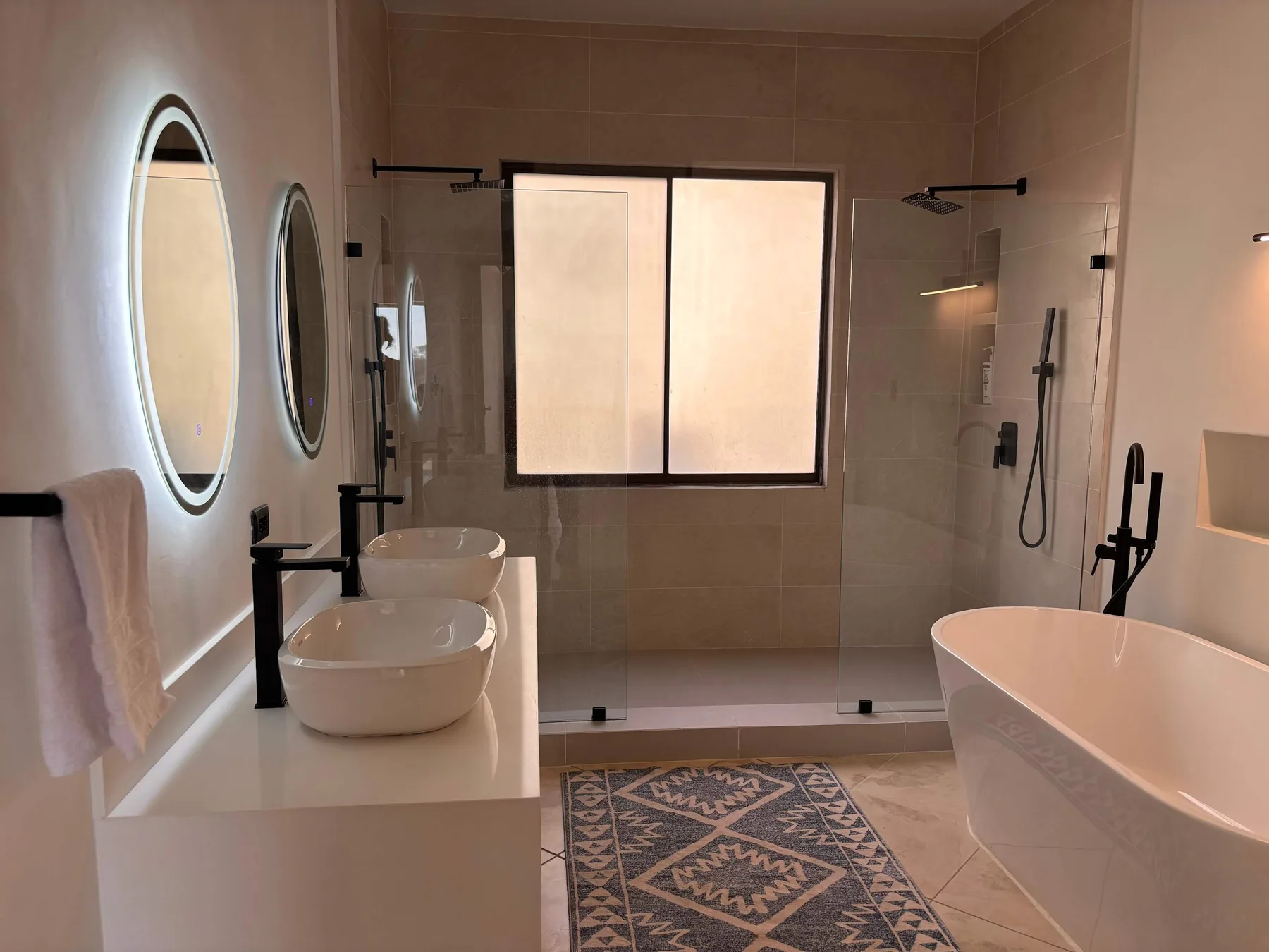 A tiled bathroom with a bathtub, two sinks, and a window.