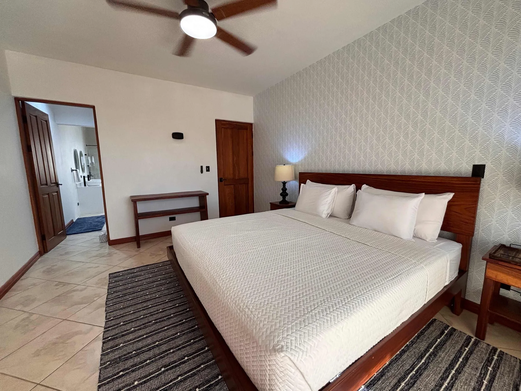 A bedroom with a large bed, patterned wallpaper, and a ceiling fan.