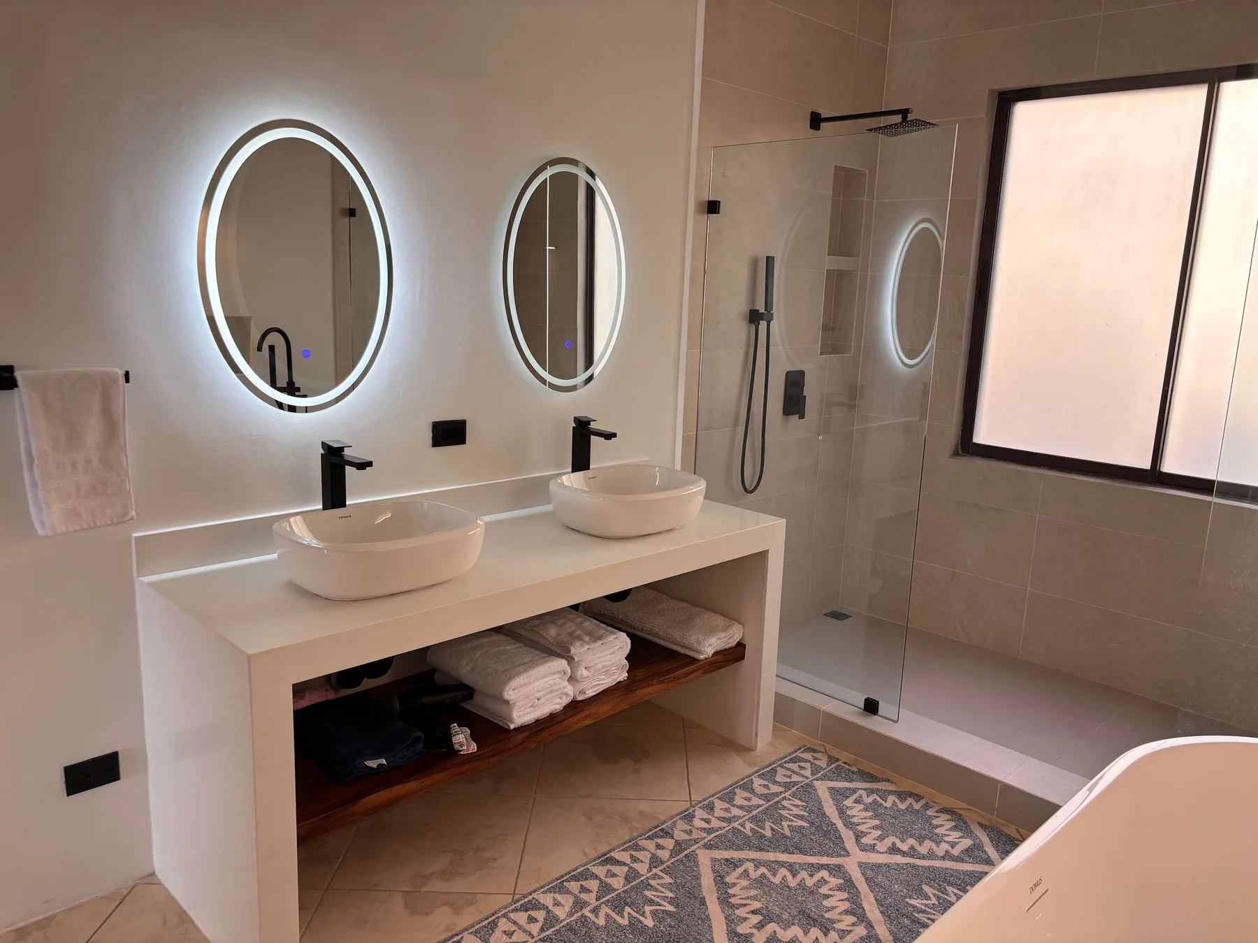 A bathroom vanity with two sinks and illuminated mirrors beside a glass shower.