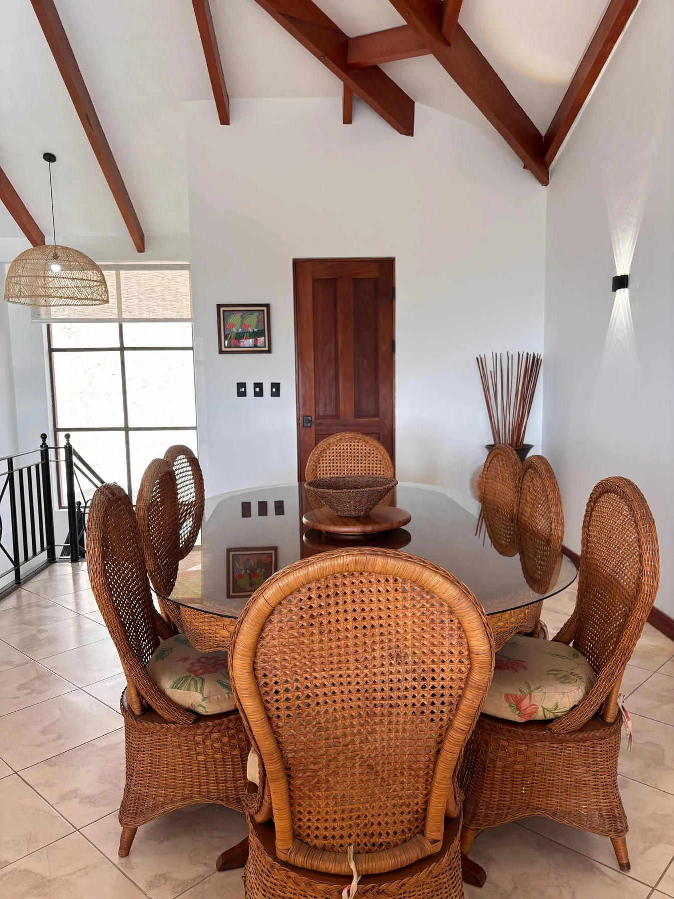 A dining area with a glass table surrounded by woven seating beneath exposed wooden beams.