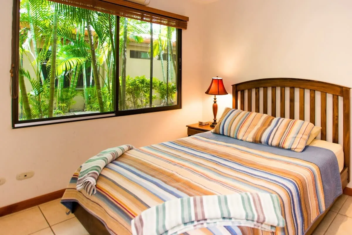 Un dormitorio con una cama a rayas junto a una ventana grande con vistas a la vegetación.