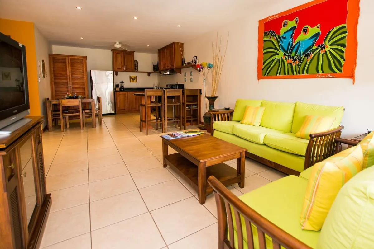 Una sala de estar con asientos verdes y una mesa de centro de madera junto a una cocina abierta.
