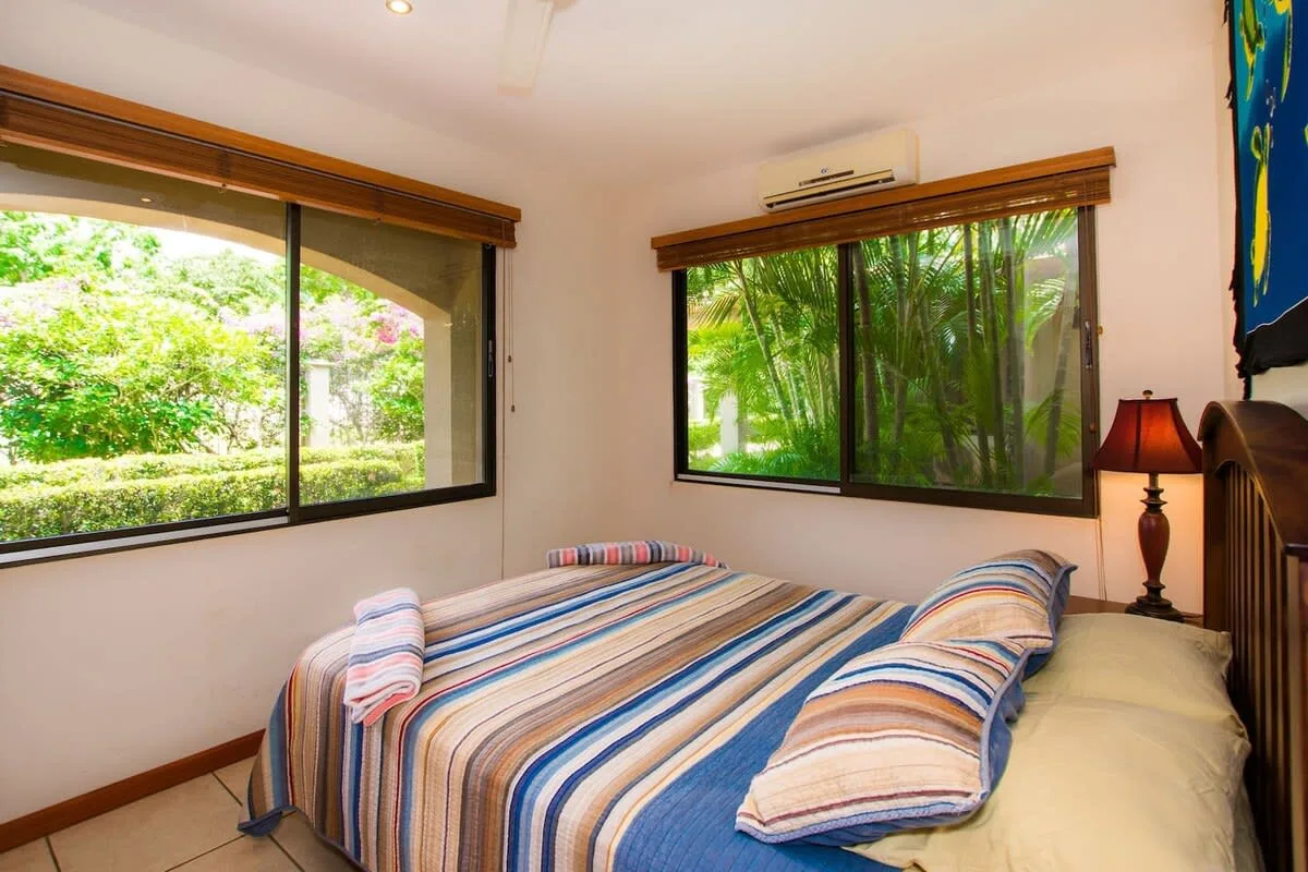 Un dormitorio con una colcha a rayas y grandes ventanas con vistas a la vegetación.