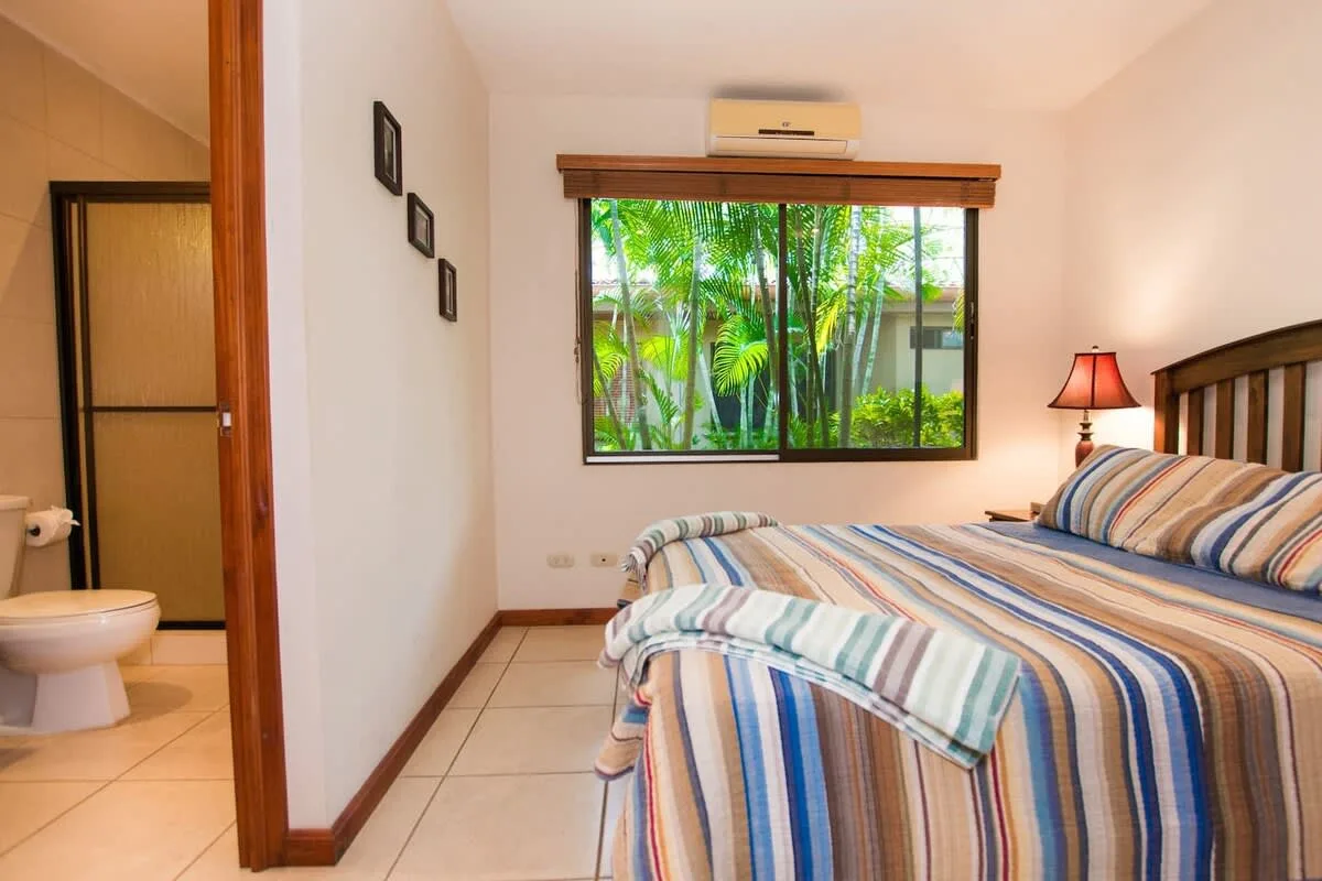 Un dormitorio con una cama a rayas junto a una ventana con vegetación.