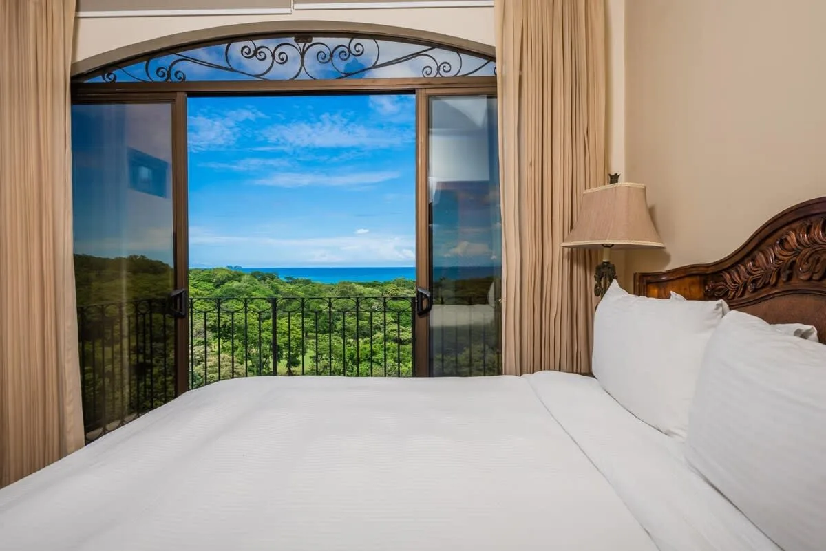 A bedroom opens onto a balcony with views of green foliage and the sea.