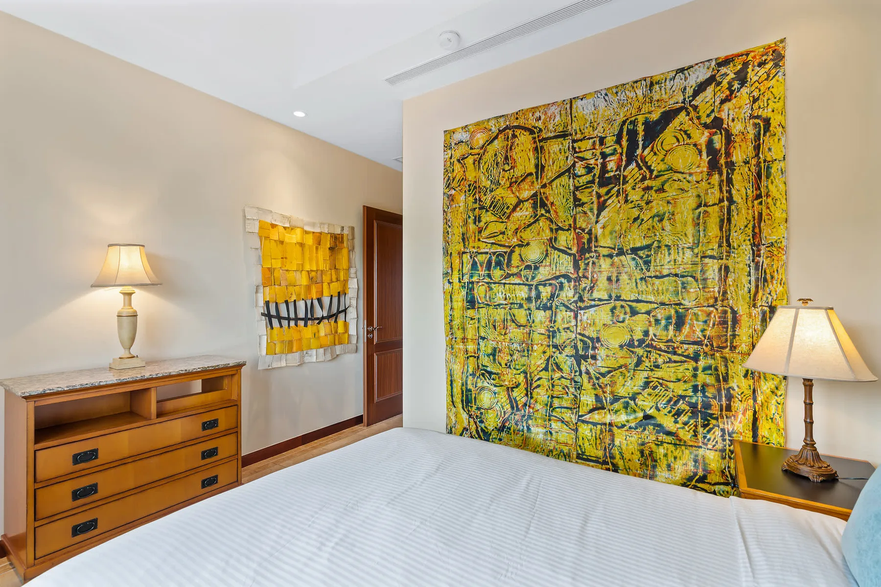 A bedroom with a bed, wooden dresser, and colorful abstract wall art.