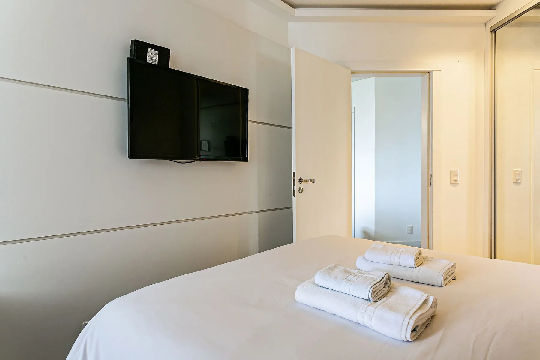 A bedroom with a wall-mounted television and folded towels on the bed.