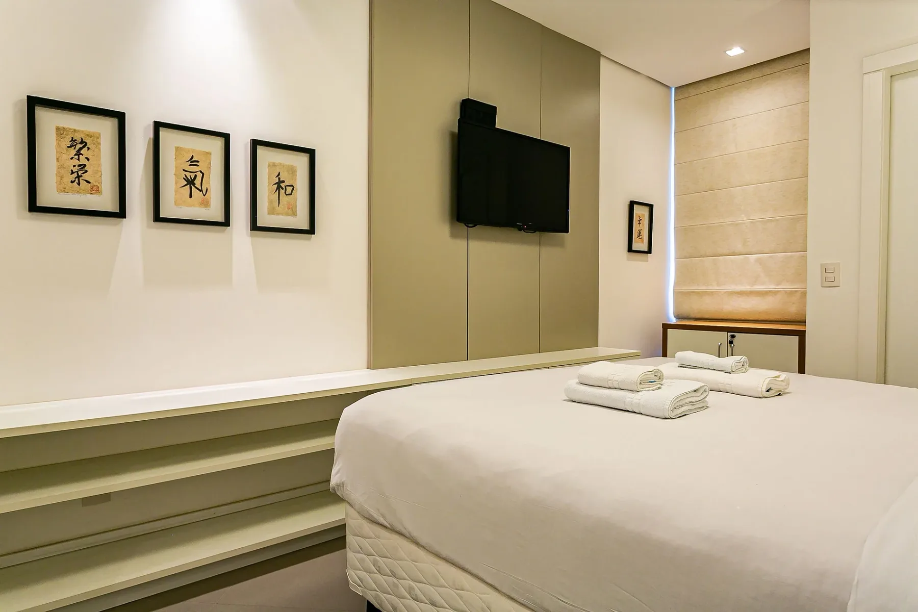 A bedroom with a neatly made bed, framed artwork, and a wall-mounted television.