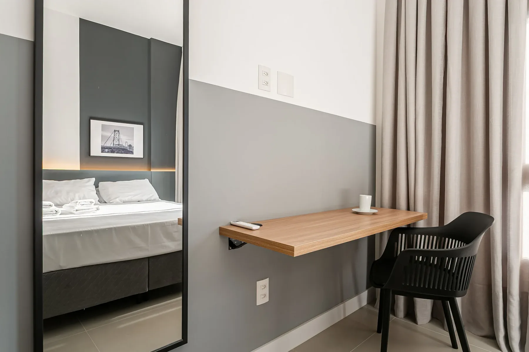 A bedroom with a bed reflected in a mirror and a desk beside a chair.