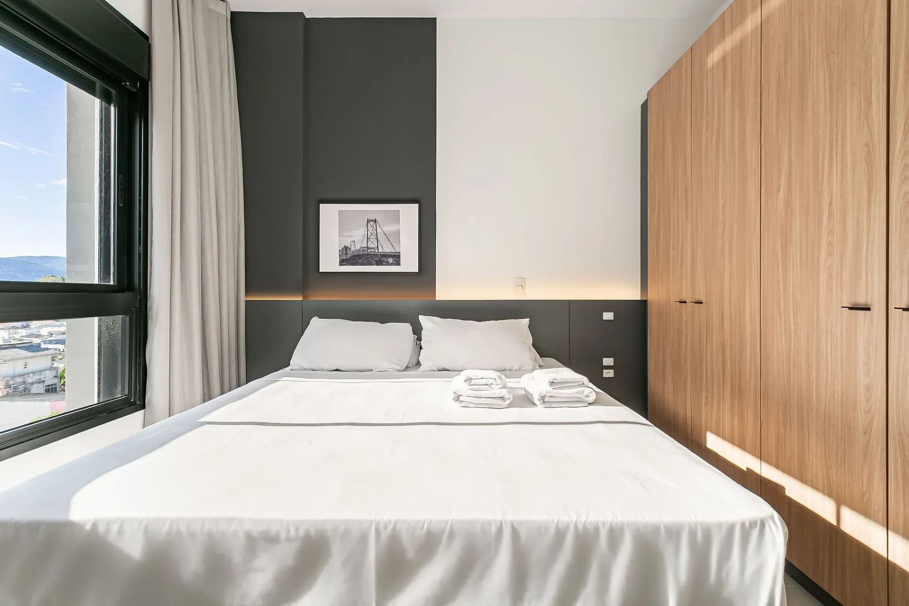 A bedroom with a neatly made bed beside a large window and wooden wardrobes.