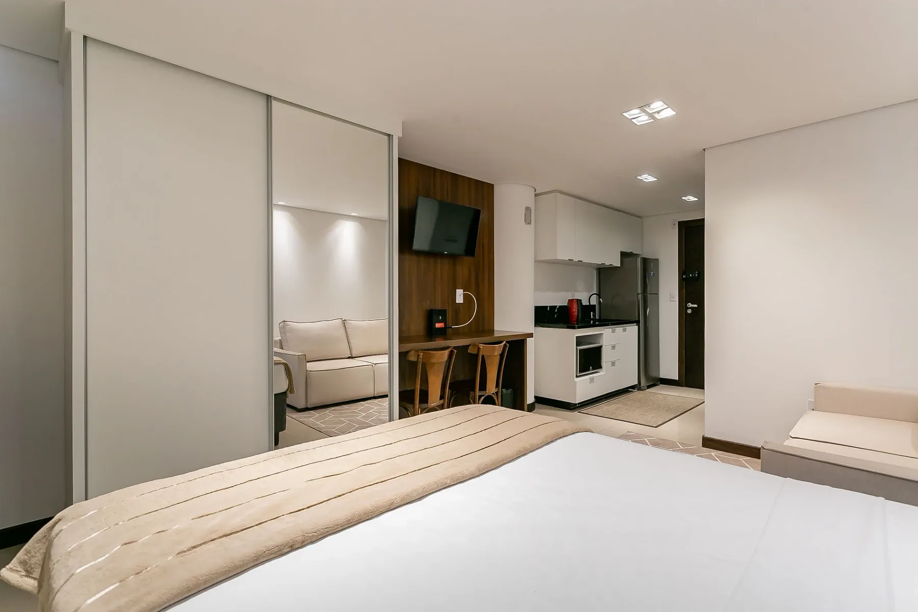 A bedroom with a bed, mirrored closet doors, and a compact kitchen area.